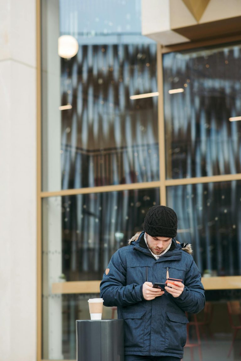 Busy young male in warm casual clothes using mobile phone while paying online for purchase with credit card standing on city street near modern building with coffee to go