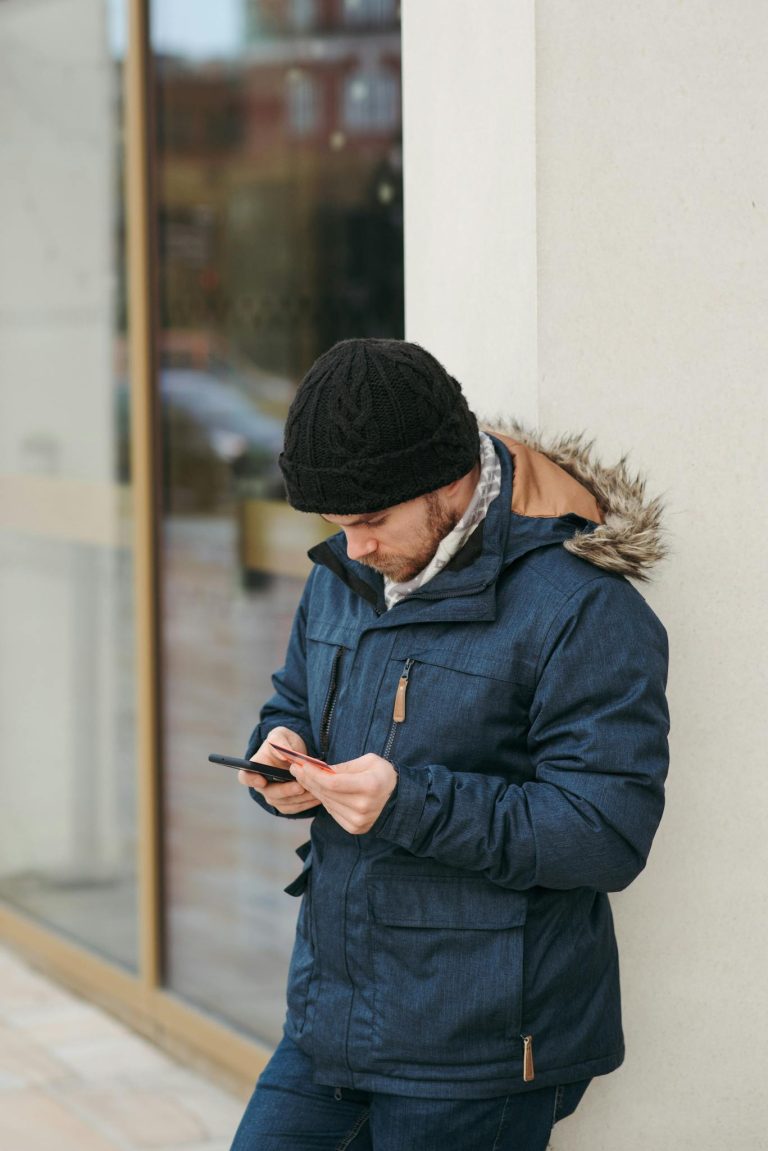 Side view of serious young male in warm clothes and hat standing on city street and using mobile phone while making online payment via credit card